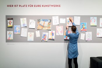 eine Besucherin pinnt ein von Kindern gezeichntes Gemälde an die mit vielen weiteren Bildern behängte Magnetwand