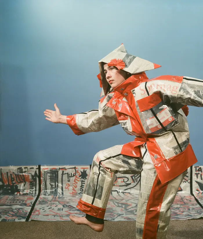 Monika Andres, Name, Stadt, Land (Zeitungskostüm und Transparent), 1988 Frau in einem Kostüm aus Zeitungspapier vor blauem Hintergrund