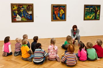 Kinder sitzen vor Gemälden, Pädagogin erzählt dazu