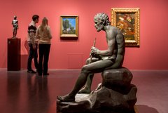 die Skulptur eines sitzenden Mannes mit Flöte, im Hintergrund Besucher, die Gemälde betrachten